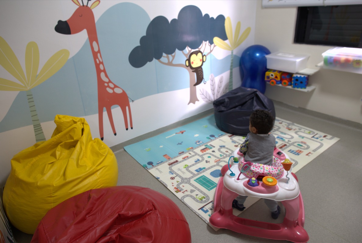 A foto mostra uma criança de costas; ela está em um andador em uma sala repleta de brinquedos e com as paredes decoradas com motivos infantis